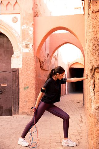 pure by luce Alou Marrakech leggings: all-over printed dark red leggings inspired by Morocco in collaboration with Hasna Lahmini and the Yattou Black sports T-shirt with cut-out sleeves worn by a girl stretching her legs against a wall in Morocco