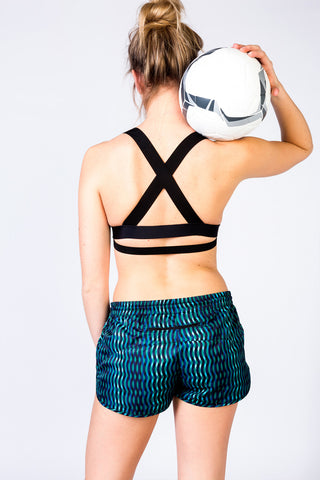 pure by luce sustainable blue all-over printed loose-fit Shourouck Akchour running shorts inspired by Morocco with the Charo Black sports bra top with cross back holding a soccer ball on her shoulder