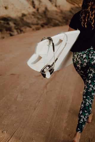 pure by luce sustainable green all-over printed Naomi Joburg leggings inspired by South Africa worn by a girl at the beach carrying a surfboard under her arm