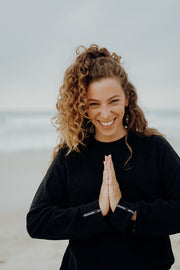 pure by luce sustainable athleisure Nina Black sweater with cut our sleeve cuffs and logo detail worn by a woman with long curly brown hair smiling and with her hands in praying position