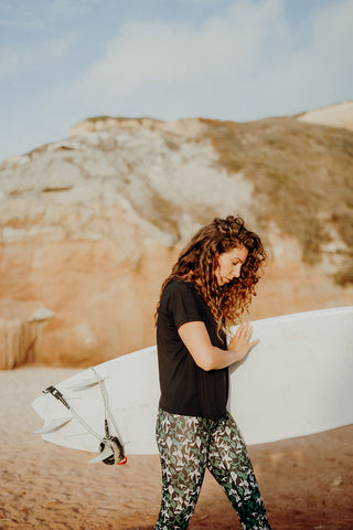 pure by luce sustainable Susan Black sports T-shirt with open back with all-over printed Naomi Joburg leggings worn by a woman with long curly brown hair carrying a surfboard under her arm