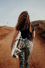 pure by luce sustainable Susan Black sports T-shirt with open back with all-over printed Naomi Joburg leggings worn by a woman with long curly brown hair carrying a surfboard under her arm